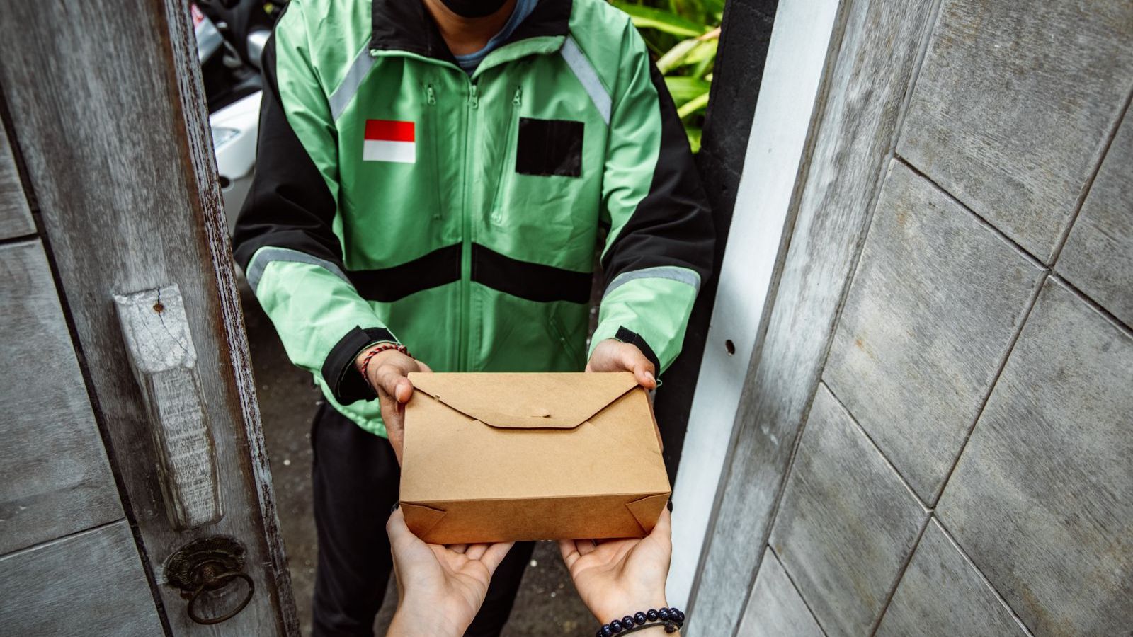 Delivery rider in a green jacket handing a parcel to a customer at the door, representing proof of delivery